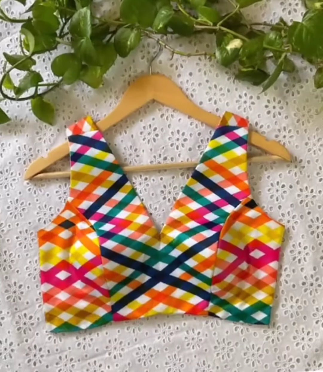 Colourful geometric-patterned blouse on a wooden hanger against a white lace background with green leaves.