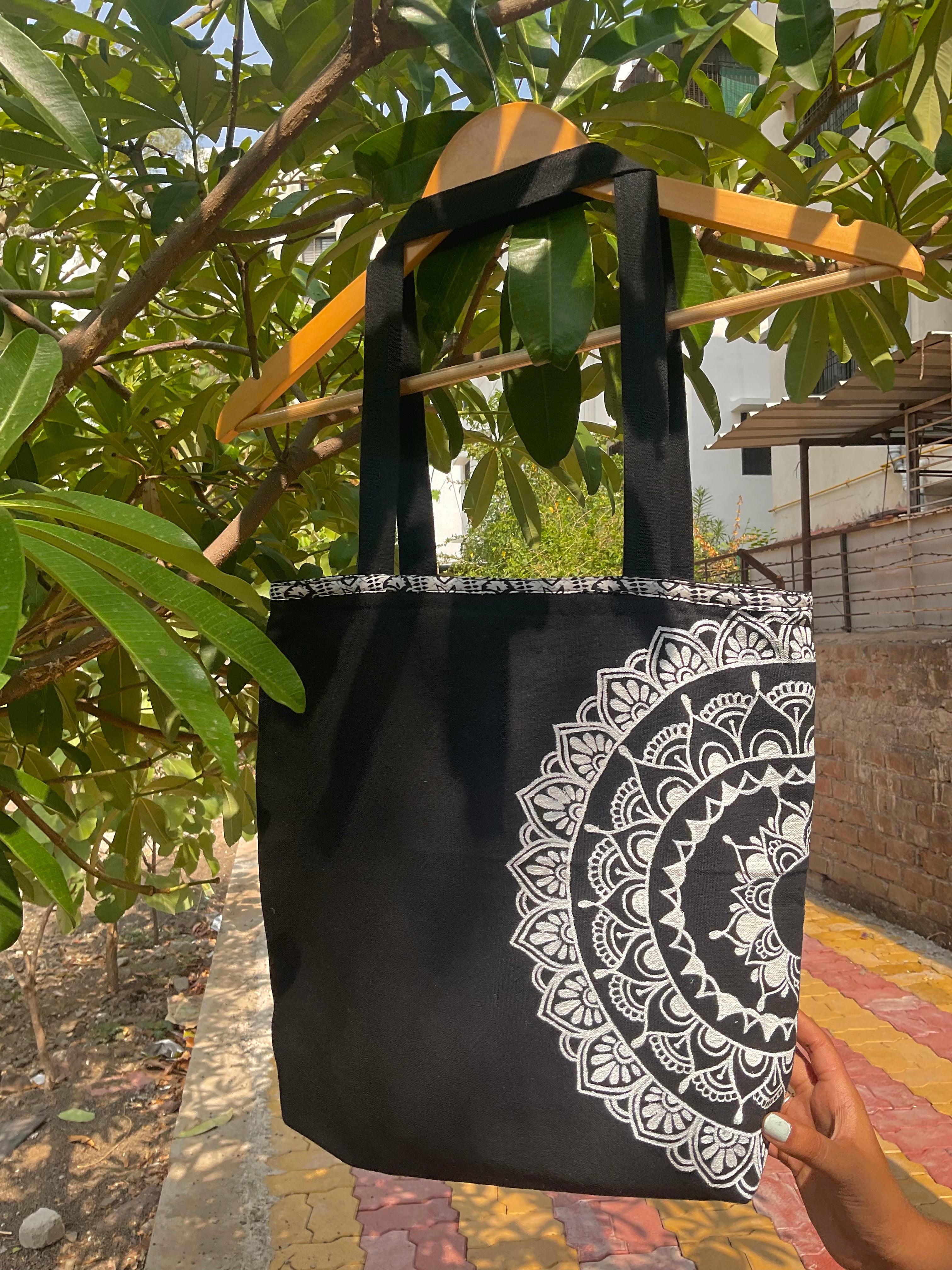 Black tote bag with white mandala design held by a person outdoors.