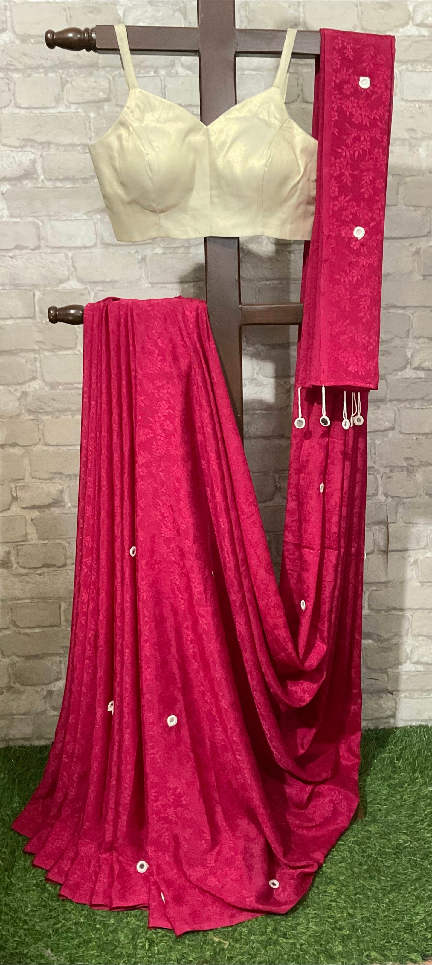 Beige blouse and pink saree with a wooden rod against a brick wall.