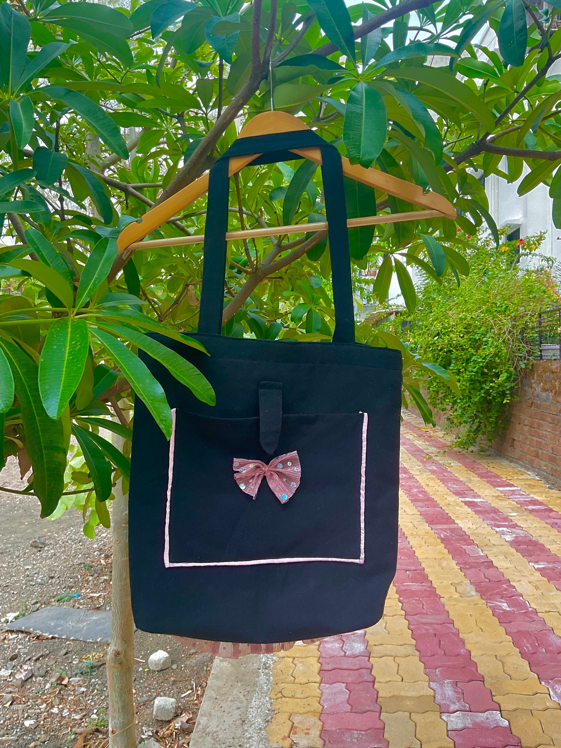 Black tote bag with a pink bow on a hanger against a green leafy background