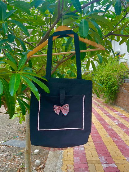 Black tote bag with a pink bow on a hanger against a green leafy background