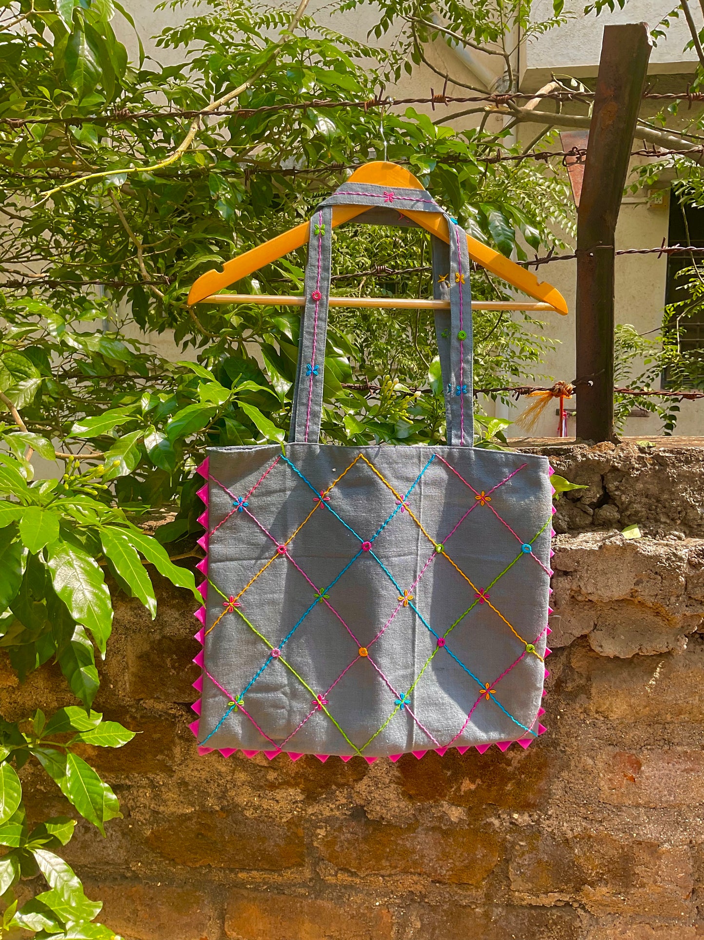 Gray tote bag with colourful patterns and triangle details hanging on a wooden stick against a stone wall with greenery.
