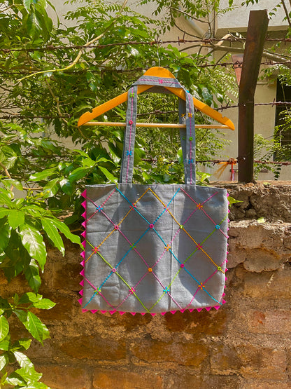 Gray tote bag with colourful patterns and triangle details hanging on a wooden stick against a stone wall with greenery.