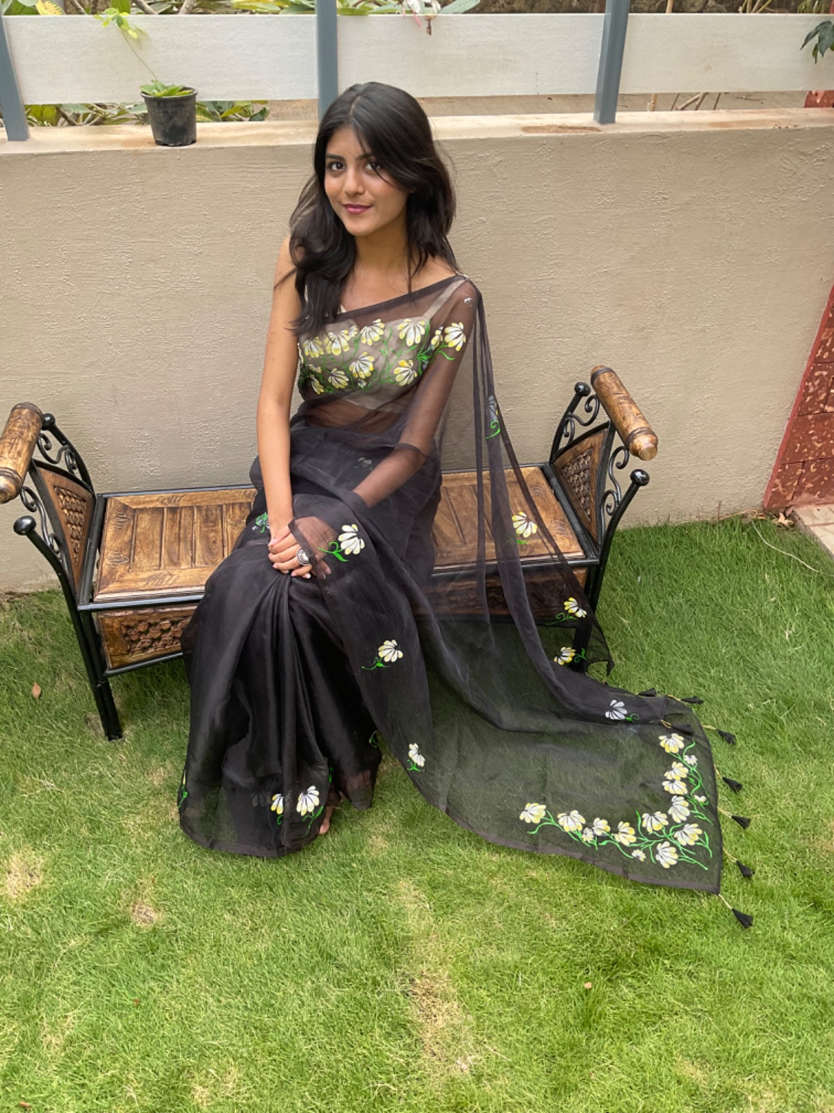 Woman in a black saree with floral patterns sitting on a wooden bench outdoors.