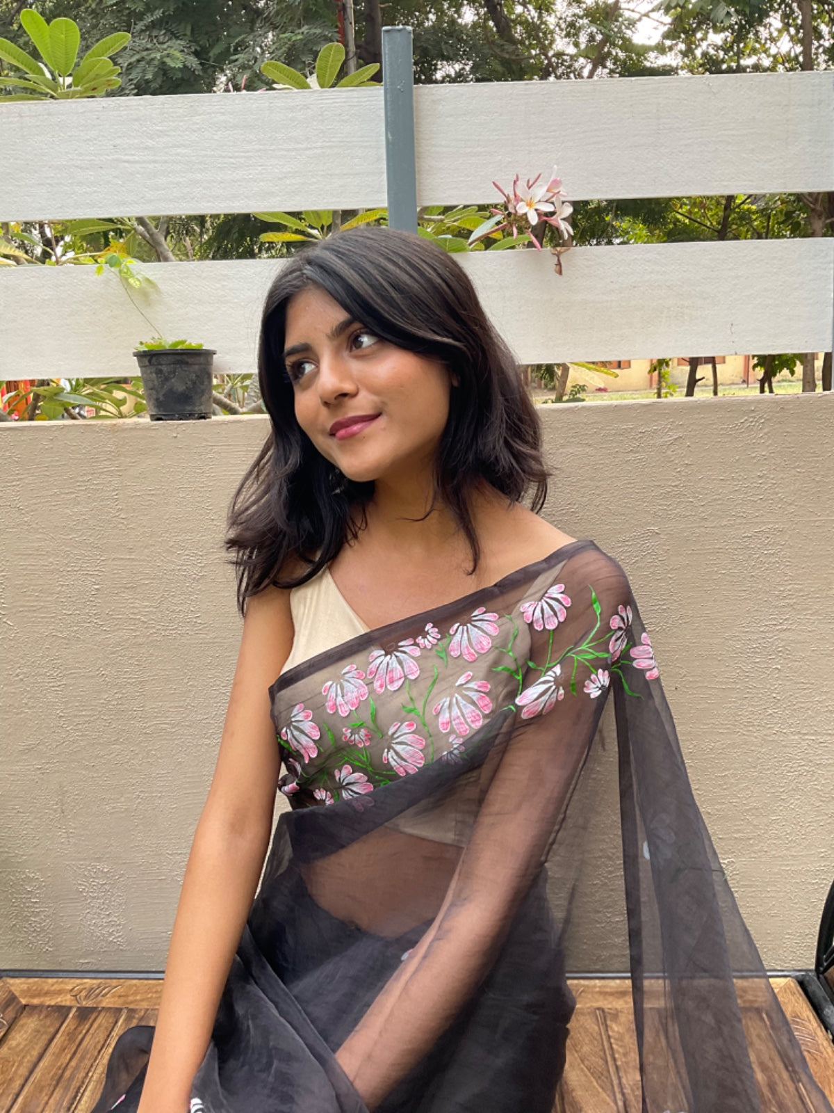 Woman wearing a black saree with floral patterns sitting outdoors.