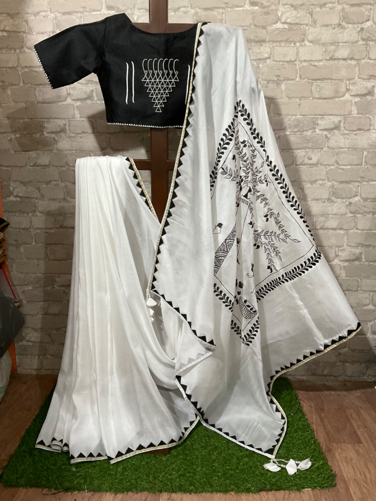 White saree with black hand painting on a green mat against a brick wall.