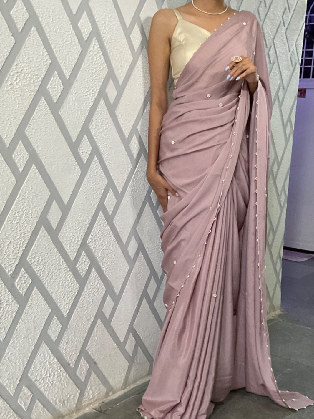Person wearing a mauve saree with a white blouse against a geometric-patterned wall.
