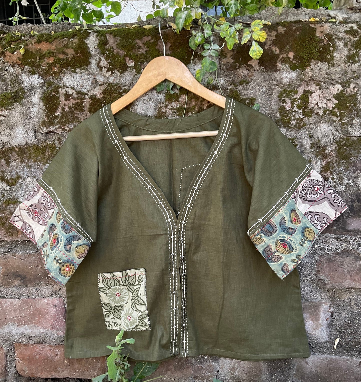 Green jacket with floral patterns on a wooden hanger against a stone wall.