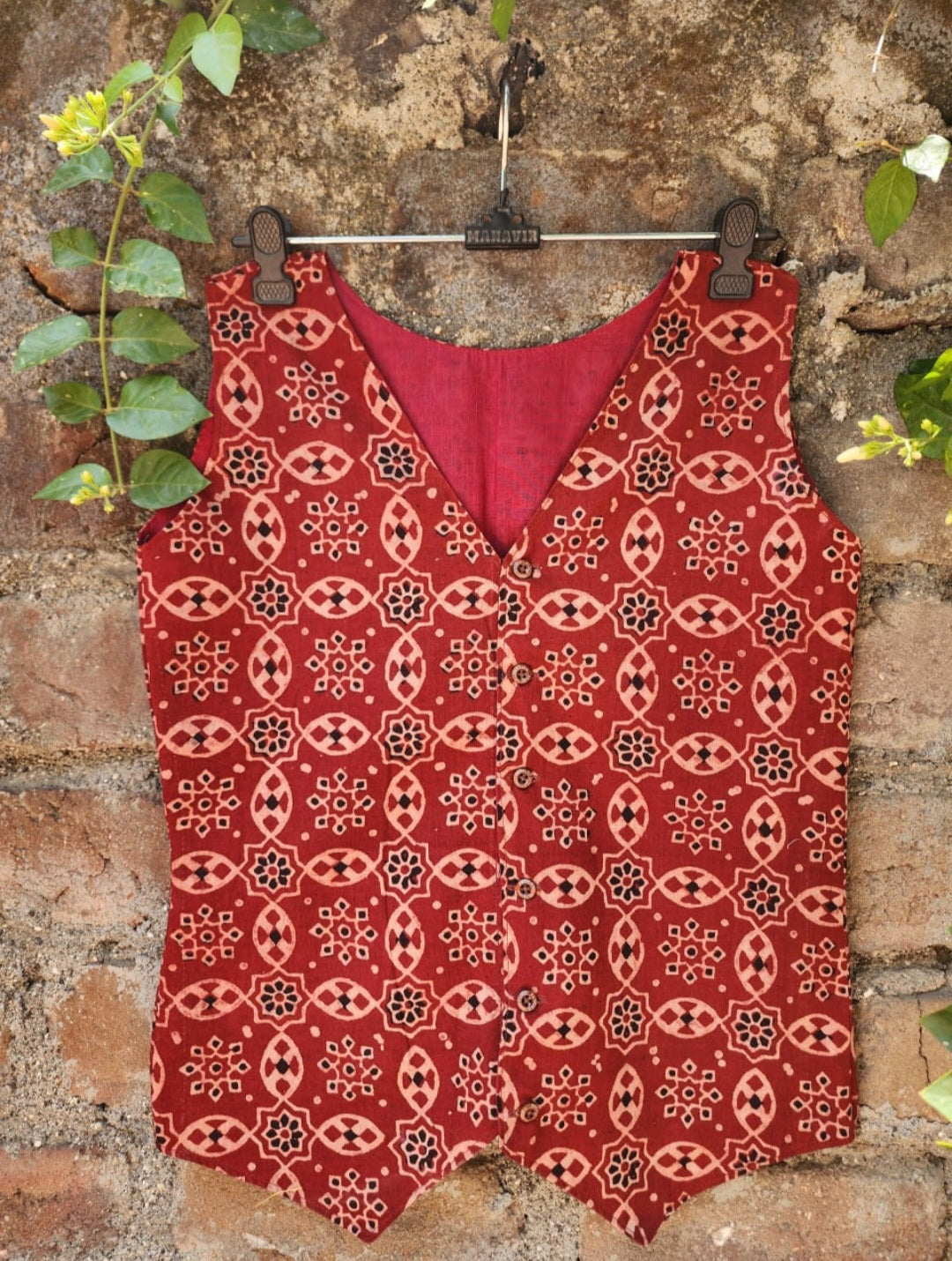 Red kalamkari vest hanging on a brick wall with green leaves.